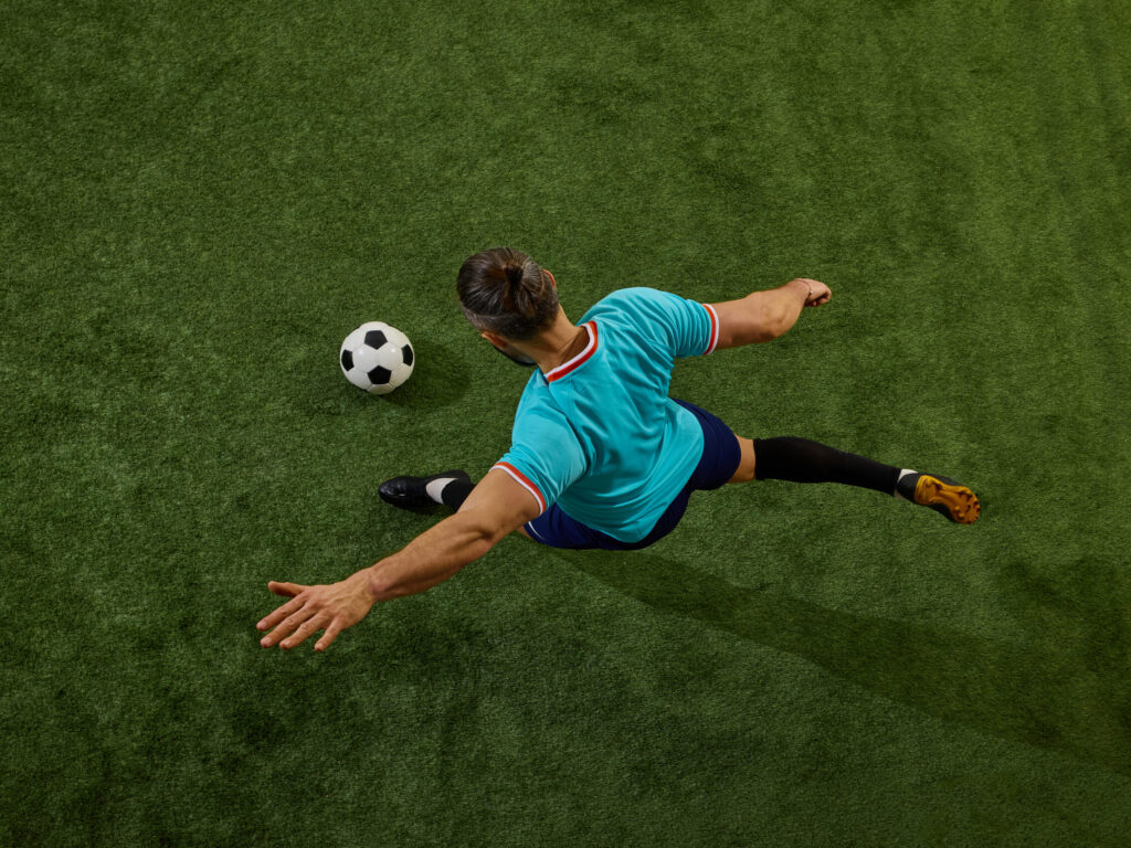 Top-down view of a soccer player in a turquoise jersey kicking a black-and-white ball on green turf.