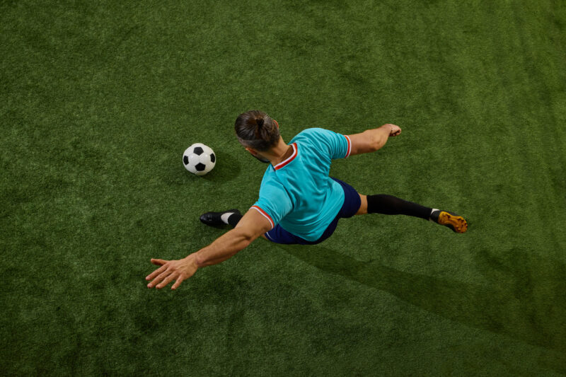 Top-down view of a soccer player in a turquoise jersey kicking a black-and-white ball on green turf.