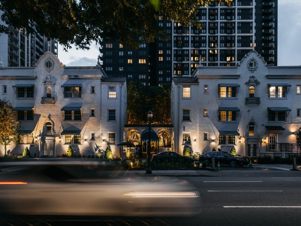 White ornate townhouses at dusk with a tall modern apartment building behind them, and light-trail cars speeding by on the street.