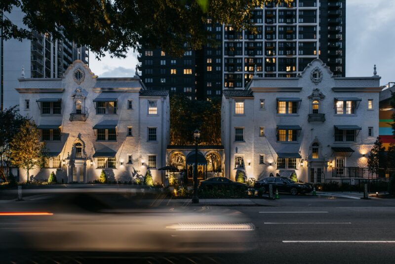 White ornate townhouses at dusk with a tall modern apartment building behind them, and light-trail cars speeding by on the street.