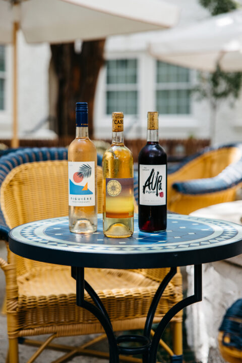 Three glass bottles on a blue patterned table outdoors, with wicker chairs in the background.