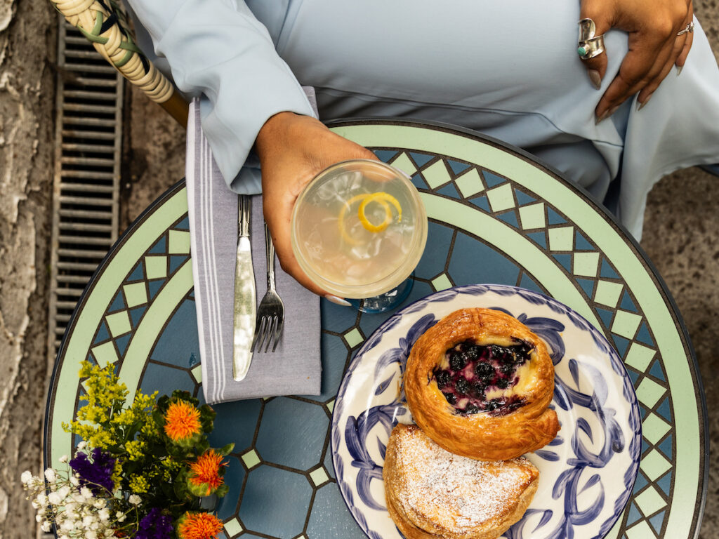 Top-down view of two people at a decorative blue mosaic table: pastries on a plate, a lemon drink, a latte with latte art, a small bouquet of flowers, and hands resting on the table in light-blue clothing.