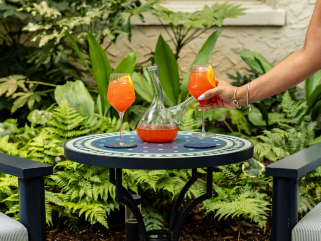 Person reaches to pick up an orange cocktail from a glass on a patterned round table in a garden setting, with a carafe nearby and two drinks total.