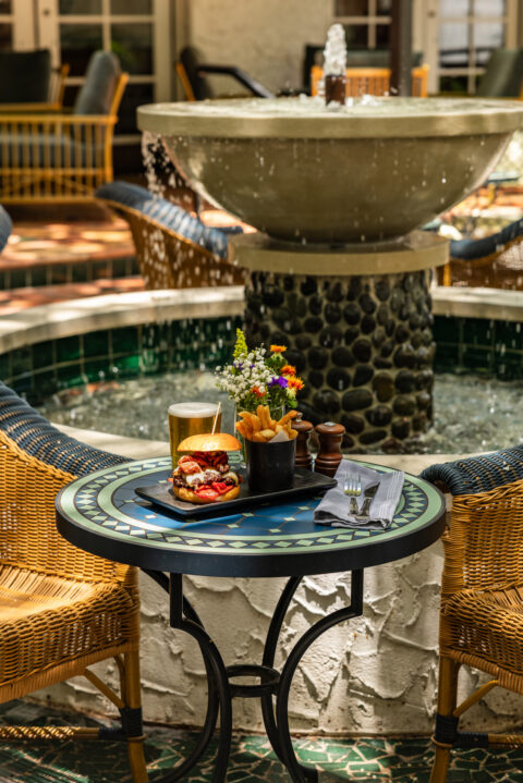 Outdoor patio scene with a burger, fries, beer, and a small bouquet, set at a decorative tiled table beside a water fountain and wicker chairs.