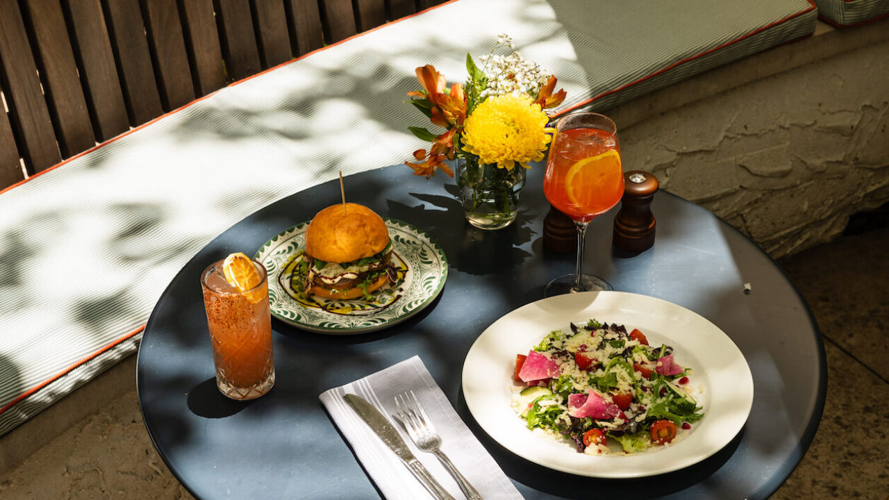 Outdoor patio table set for lunch with burger, salad, two drinks, and a yellow flower centerpiece in sunlight.