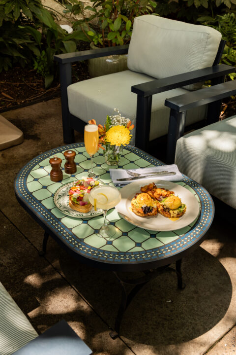 Outdoor patio scene with a mosaic glass-top table set with food and drinks; plates of sandwiches, a yellow flower bouquet, and a champagne cocktail.