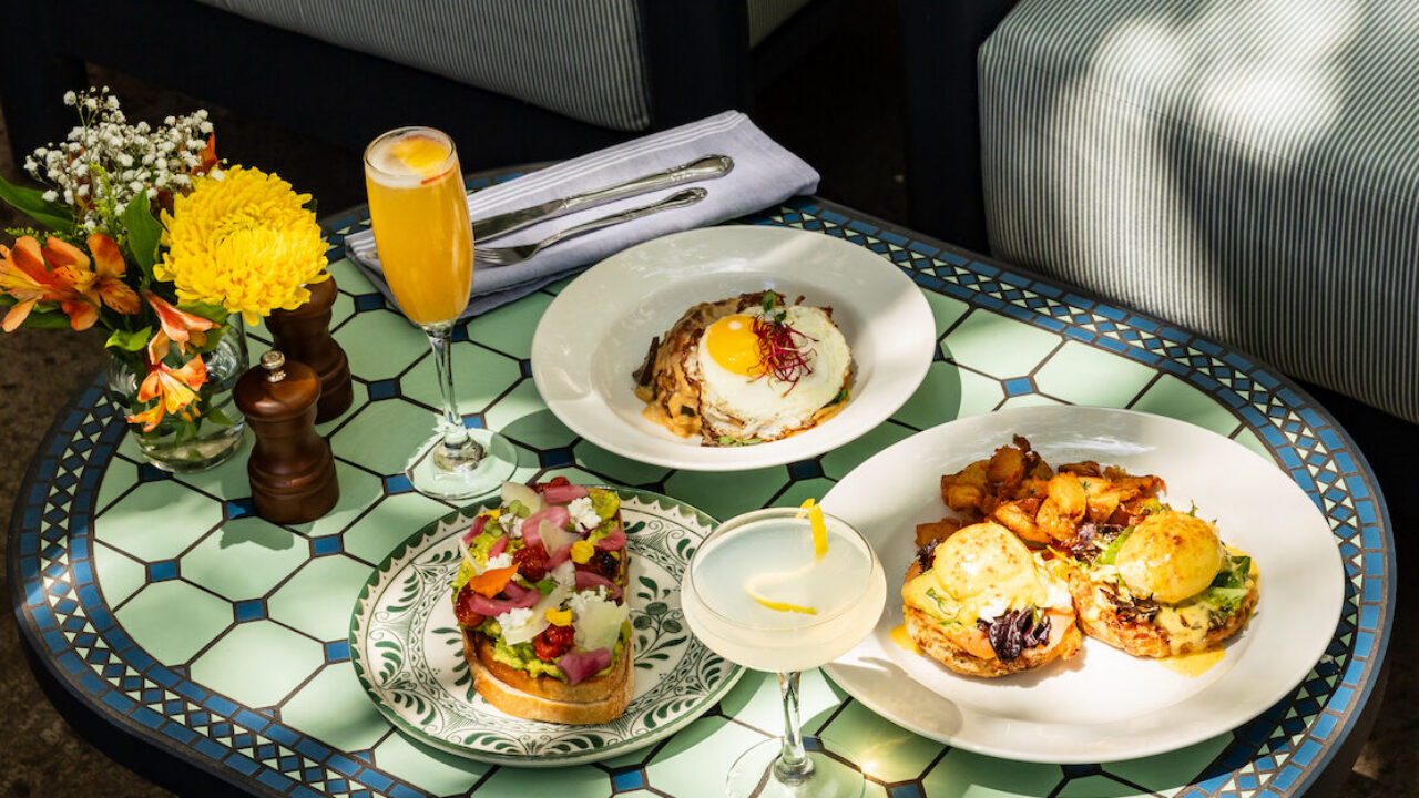 Outdoor patio scene with a mosaic-tile table holding three plated brunch dishes, a glass of orange juice, and a vase of flowers beside striped lounge chairs.